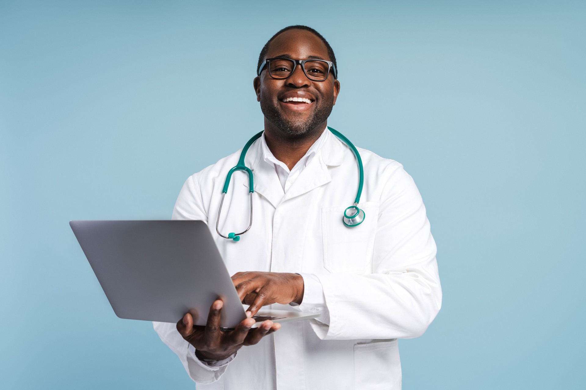 Médico afro-americano feliz em óculos segurando laptop olhando para a câmera em estúdio